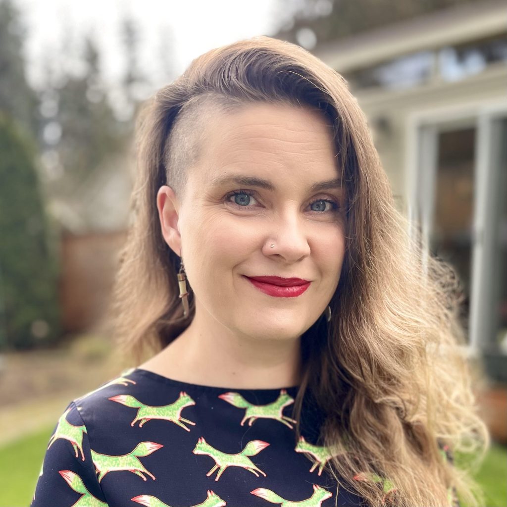 Portrait of the author, Erin M. Evans. Middle-aged White woman with long brown hair and a sideshave. She's wearing red lipstick and a shirt with green foxes on it.